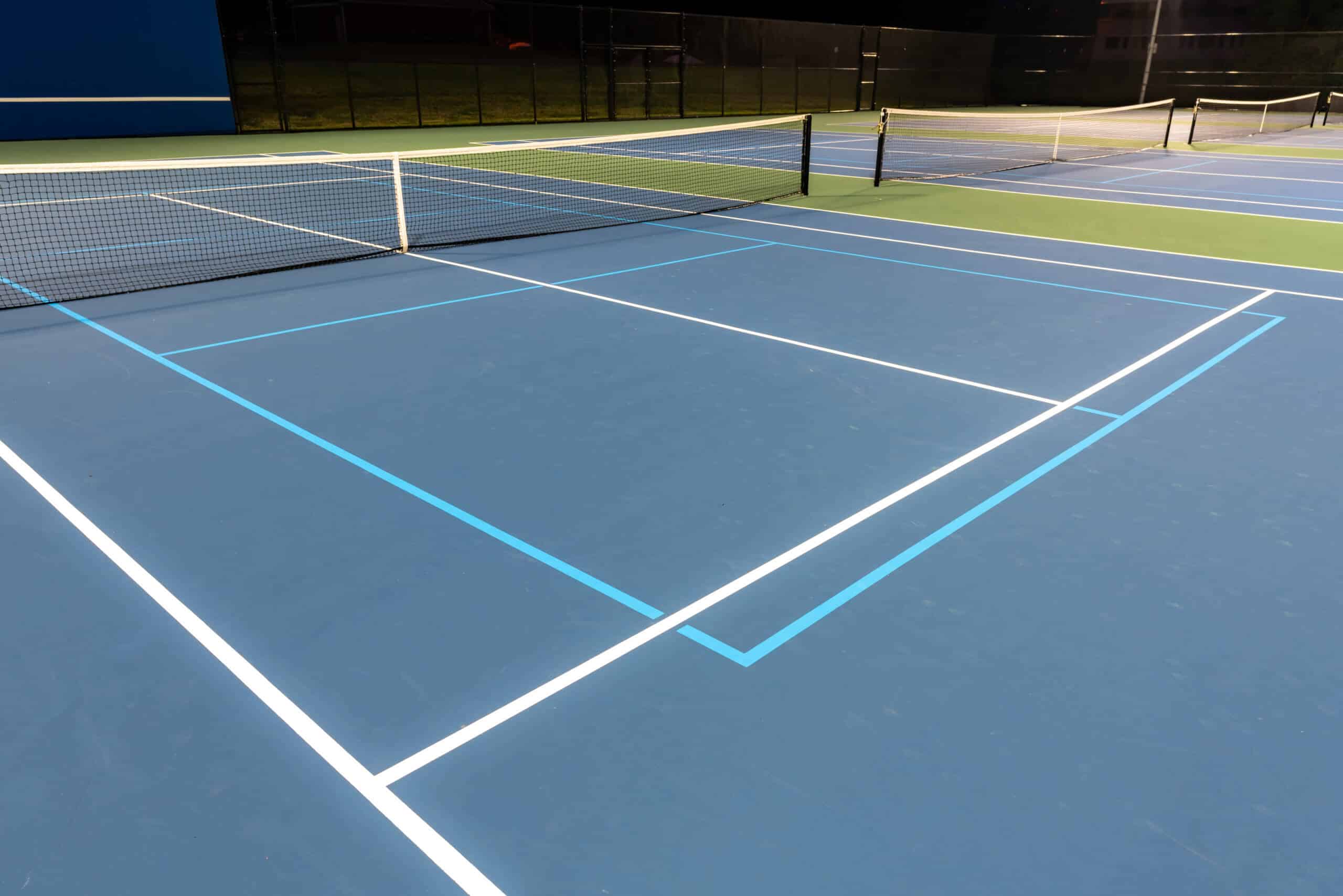 Evening photo of outdoor blue tennis courts with pickleball lines with lights turned on.