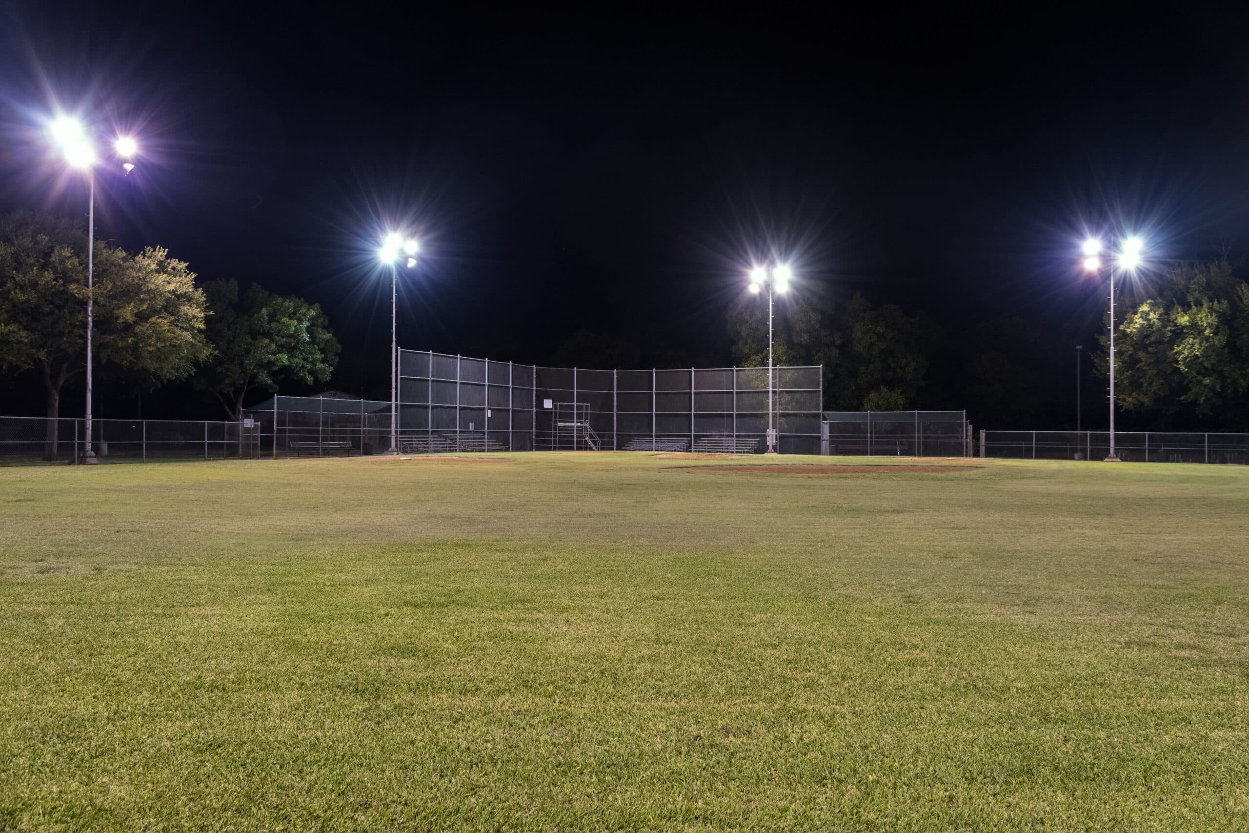 Night photo of an empty baseball field at night looking back toward home plate from right field with the lights on and contrasting against the blackness of the night sky.
