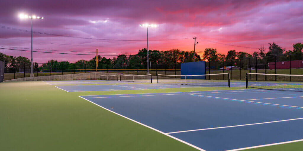 Evening photo of outdoor blue tennis courts with pickleball lines with lights turned on.